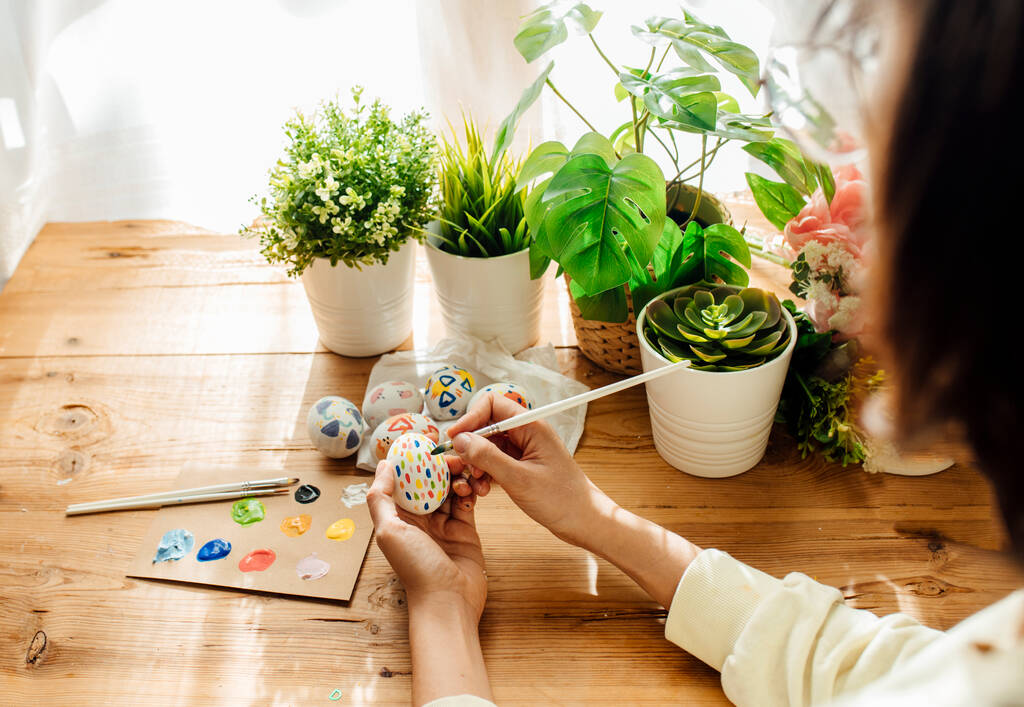 女性は手作りのイースターエッグを描いた。花や植物でブラシや塗料。木製の背景に幸せなイースターの概念. - 写真・画像