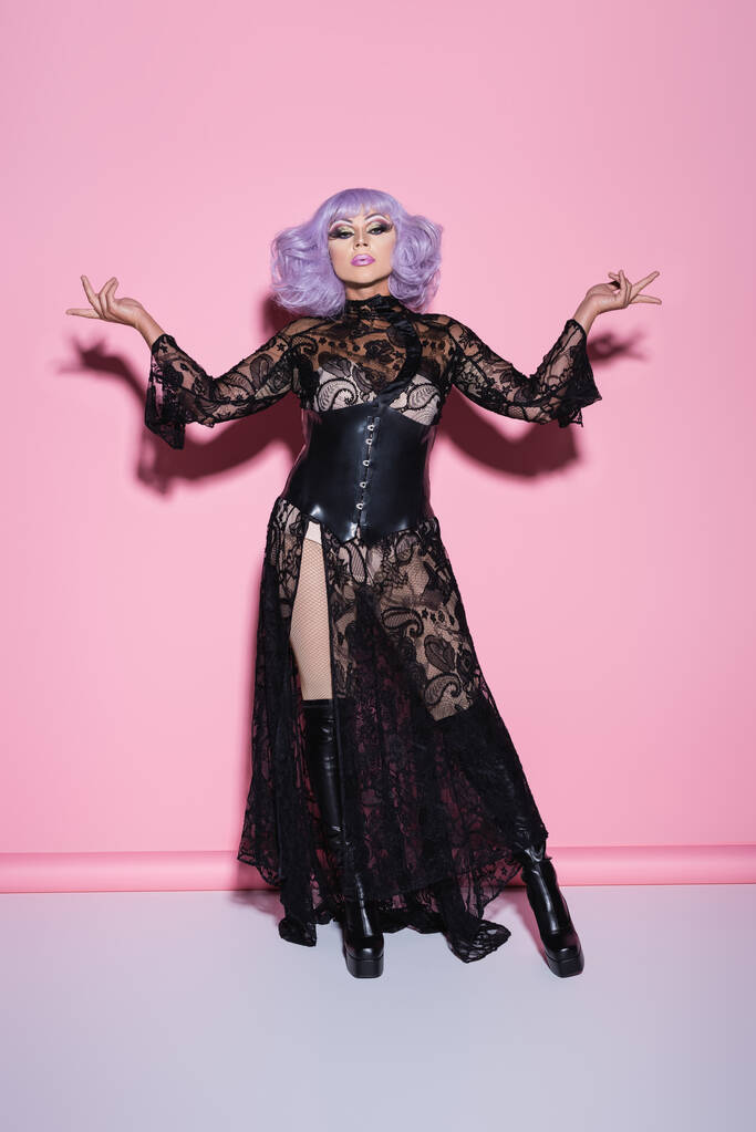 transgender in abito di pizzo nero e parrucca viola in piedi a braccia aperte su rosa - Foto, immagini
