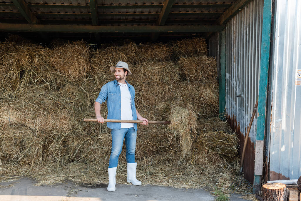 visão de comprimento total do agricultor feliz em roupas jeans e botas de borracha perto de palheiro - Foto, Imagem