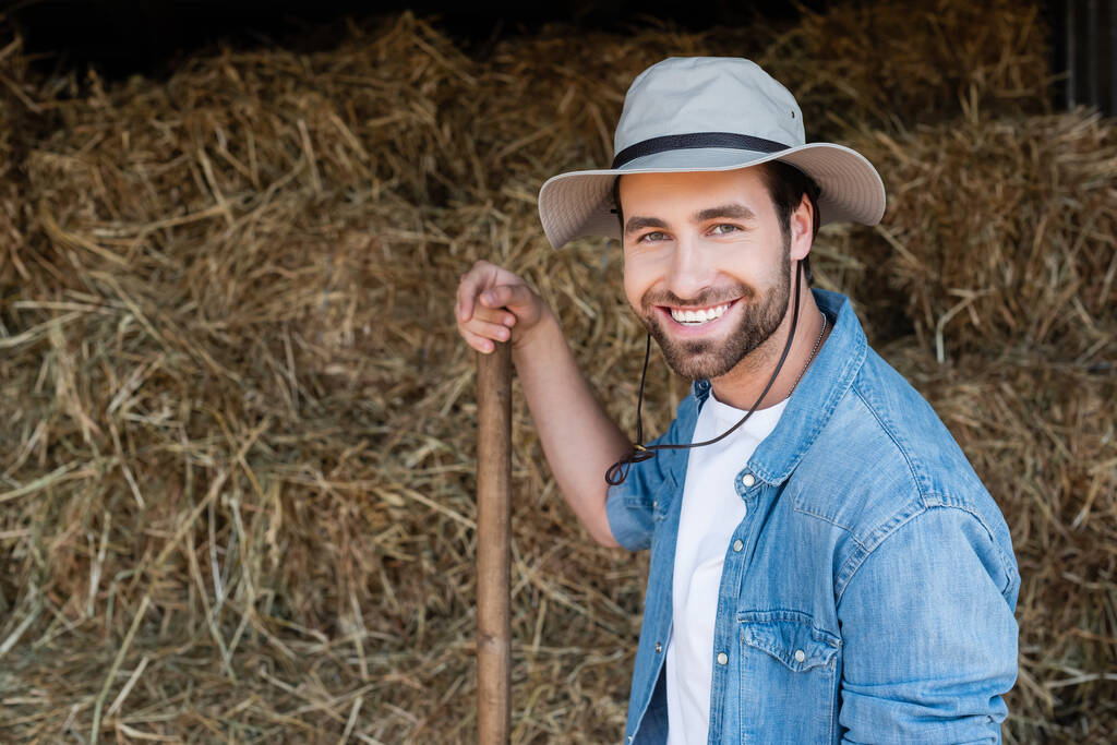 vreugdevolle boer in felle hoed kijkend naar camera in de buurt van wazig hooi op boerderij - Foto, afbeelding
