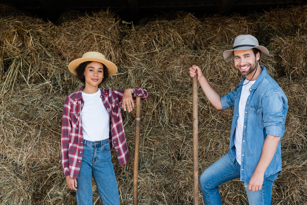 jóvenes agricultores interracial en sombreros sonriendo a la cámara cerca de pajar - Foto, Imagen