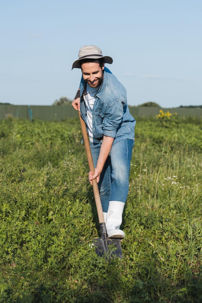 つばの帽子とデニムの服で満足した農家の完全な長さのビューフィールドで掘削 - 写真・画像