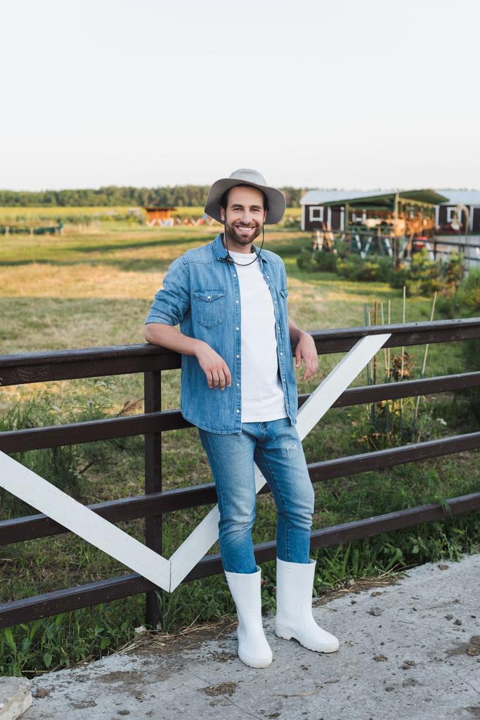 vista de longitud completa de agricultor sonriente en ropa de mezclilla de pie cerca de valla de madera en la granja - Foto, Imagen