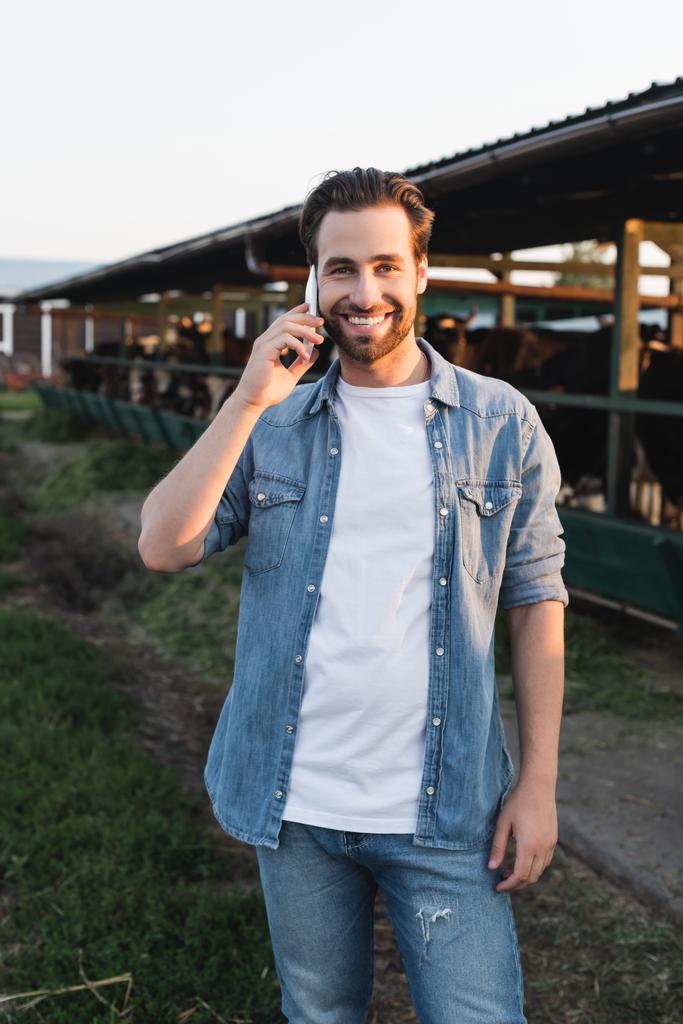 granjero hablando en el teléfono inteligente cerca borrosa vaca y sonriendo a la cámara - Foto, imagen