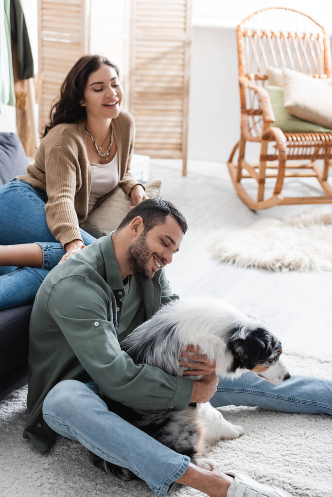 glücklicher bärtiger Mann kuschelt australischen Schäferhund neben freudiger Freundin auf Sofa - Foto, Bild