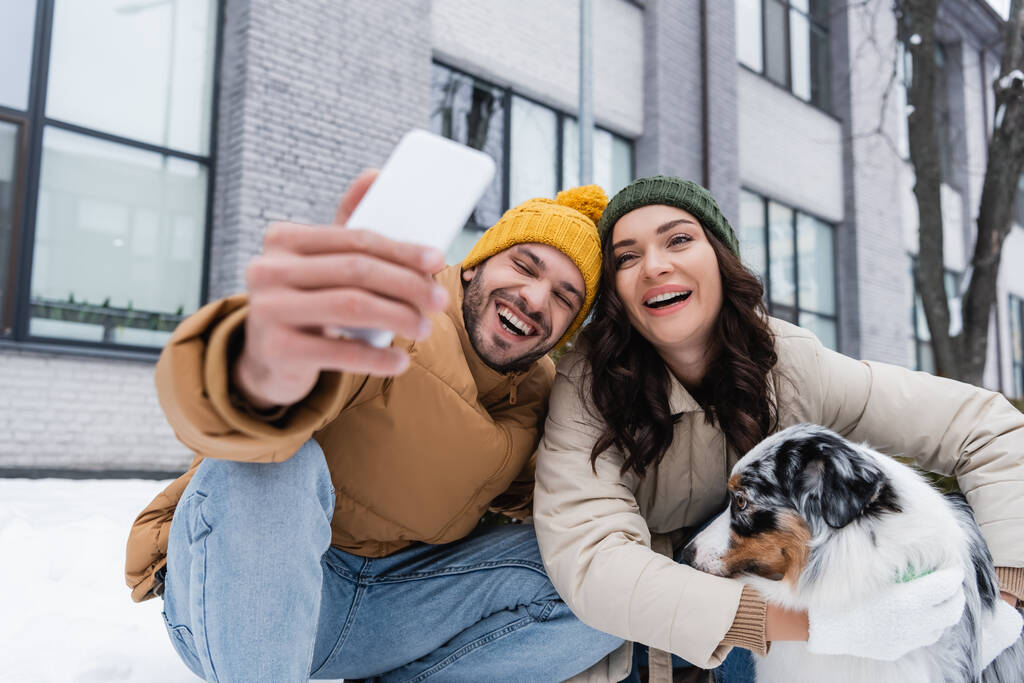 Lächelnder Mann mit Strickmütze macht Selfie mit Freundin und australischem Schäferhund im Winter  - Foto, Bild