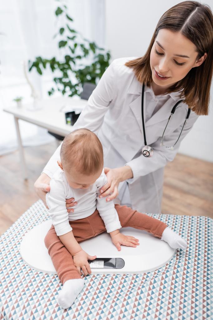 Улыбающийся врач держит ребенка на электронном весе в клинике  - Фото, изображение