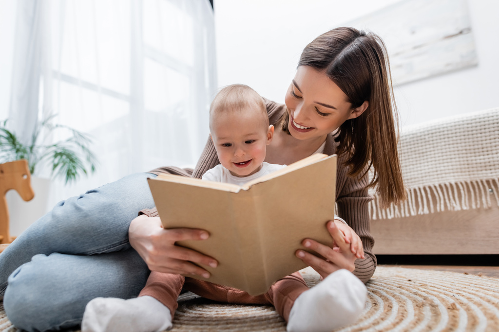 Mosolygó nő olvas elmosódott könyv közelében baba szőnyegen otthon  - Fotó, kép