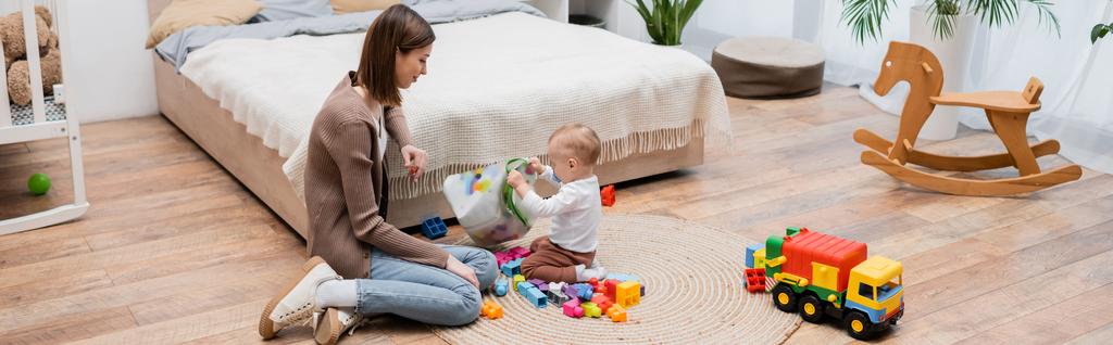 Woman looking at baby son holding bag with building blocks in bedroom, banner  - Photo, Image