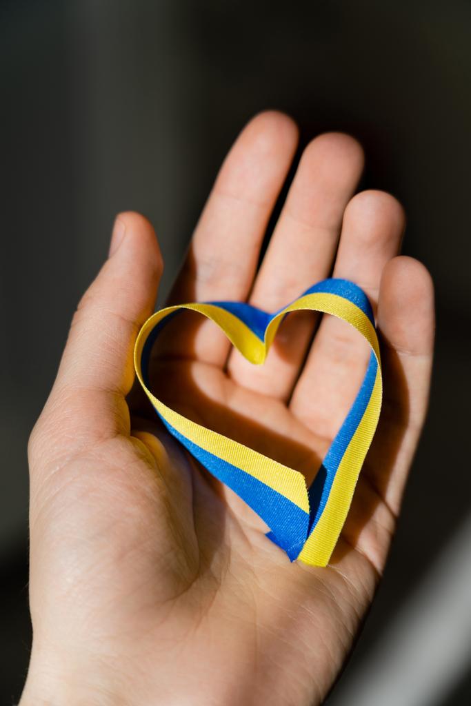 Обрізаний вид синьо-жовтої стрічки у формі серця на чоловічій руці на темно-сірому фоні
  - Фото, зображення