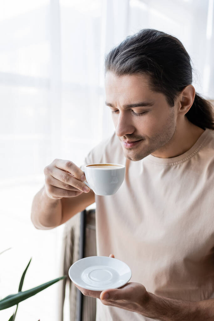 άνδρας με μακριά μαλλιά κρατώντας φλιτζάνι καφέ και πιατάκι  - Φωτογραφία, εικόνα
