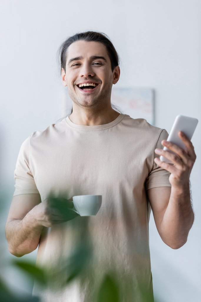 Bir fincan kahve ve akıllı telefon tutan tişörtlü mutlu bir adam. - Fotoğraf, Görsel