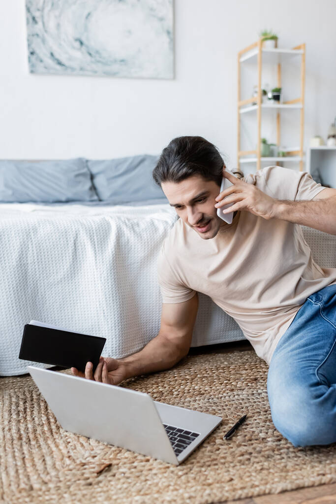 mężczyzna trzyma notatnik i rozmawia na smartfonie w pobliżu laptopa w sypialni  - Zdjęcie, obraz
