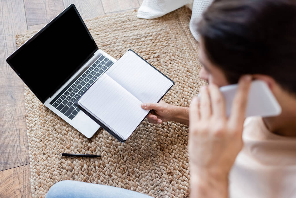 hoge hoek weergave van wazig man met notebook en praten op smartphone in de buurt van laptop met een leeg scherm  - Foto, afbeelding