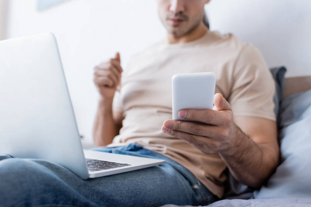 cropped view of man holding smartphone near laptop in bedroom - Photo, Image