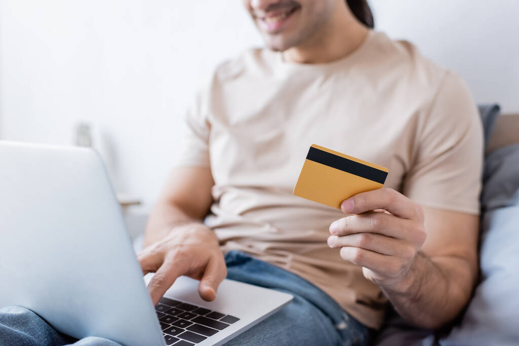 bijgesneden weergave van een gelukkige man met een creditcard en het gebruik van laptop in de slaapkamer - Foto, afbeelding