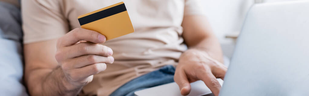 περικοπή άποψη του ανθρώπου που κατέχουν πιστωτική κάρτα και τη χρήση φορητού υπολογιστή στην κρεβατοκάμαρα, πανό - Φωτογραφία, εικόνα