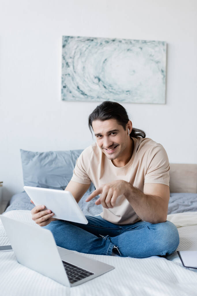 χαρούμενος άνθρωπος στο ακουστικό δείχνει σε ψηφιακή tablet κοντά στο laptop και smartphone στο κρεβάτι - Φωτογραφία, εικόνα