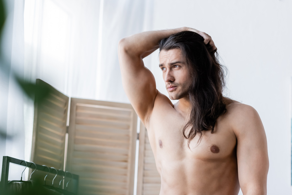 Muž bez trička, upravující si dlouhé vlasy a odvracející pohled - Fotografie, Obrázek