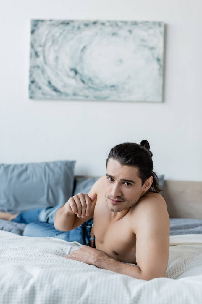 χωρίς πουκάμισο άντρας με μπλε τζιν ξαπλωμένος στο κρεβάτι και κοιτάζοντας την κάμερα - Φωτογραφία, εικόνα