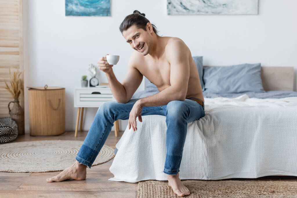 veselý muž s dlouhými vlasy drží šálek kávy, zatímco sedí na posteli - Fotografie, Obrázek