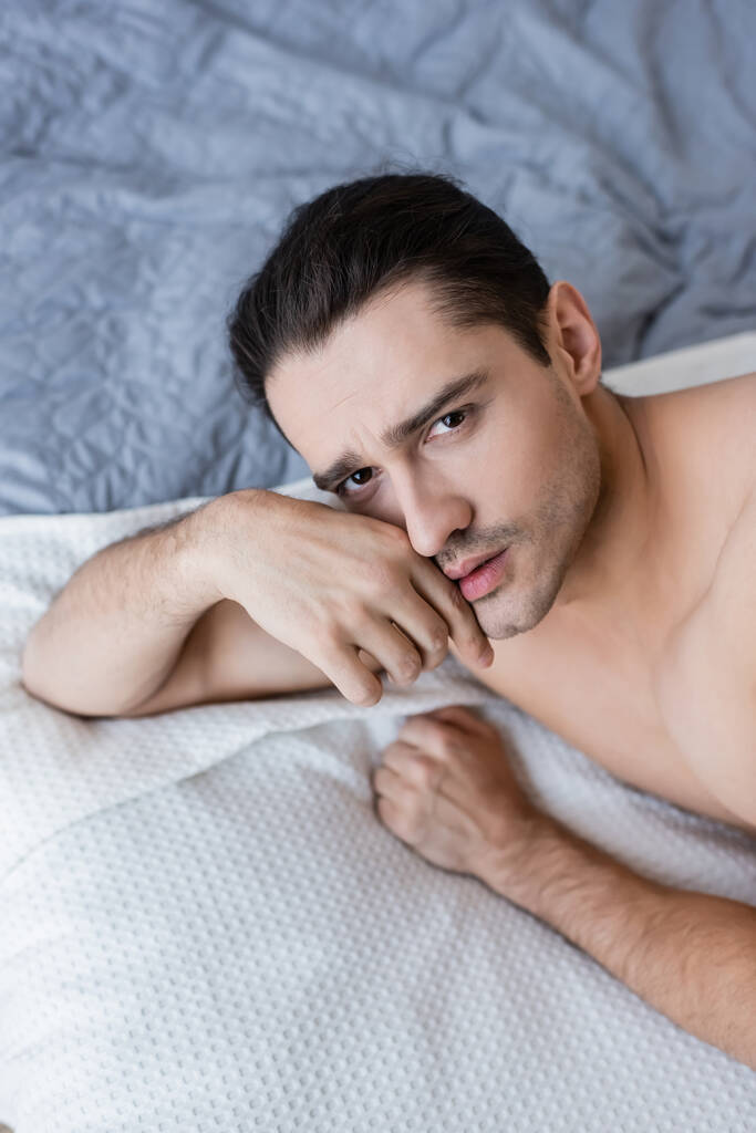 vysoký úhel pohledu na muže bez trička ležícího na posteli a dívajícího se do kamery - Fotografie, Obrázek