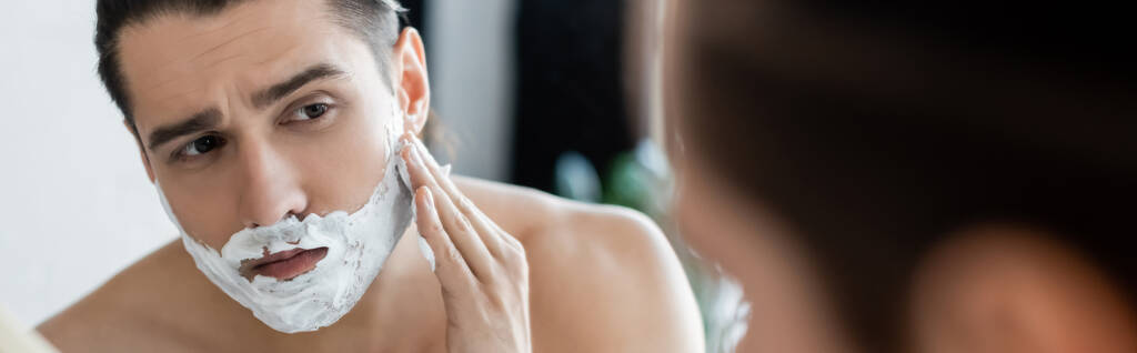 Мужчина без рубашки наносит пену для бритья на лицо и смотрит на зеркало, баннер - Фото, изображение