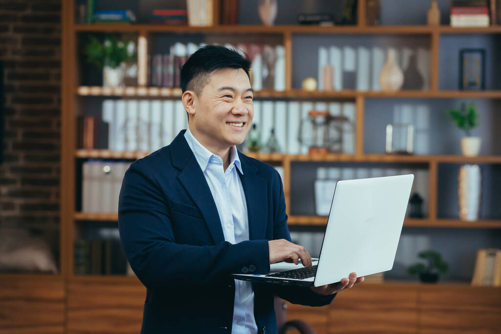 Szczęśliwy azjatycki wolny strzelec biznesmen pracujący na stojąco, trzymający laptopa, uśmiechnięty i radujący się, w biurze - Zdjęcie, obraz