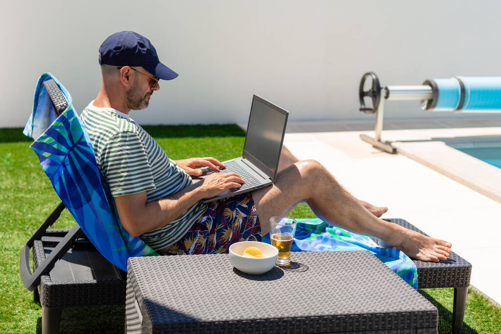 Uvolněný dospělý muž pracující na notebooku a mající aperitiv v blízkosti bazénu na letní dovolenou - Fotografie, Obrázek