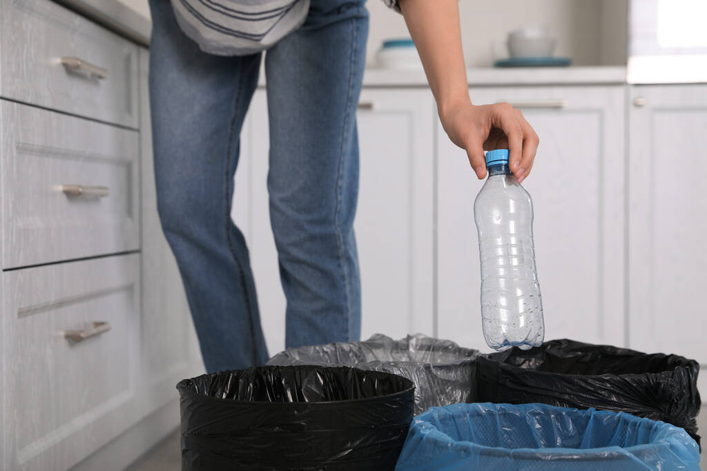 Женщина выбросила пластиковую бутылку в мусорный бак на кухне, крупным планом. Отдельный сбор отходов - Фото, изображение