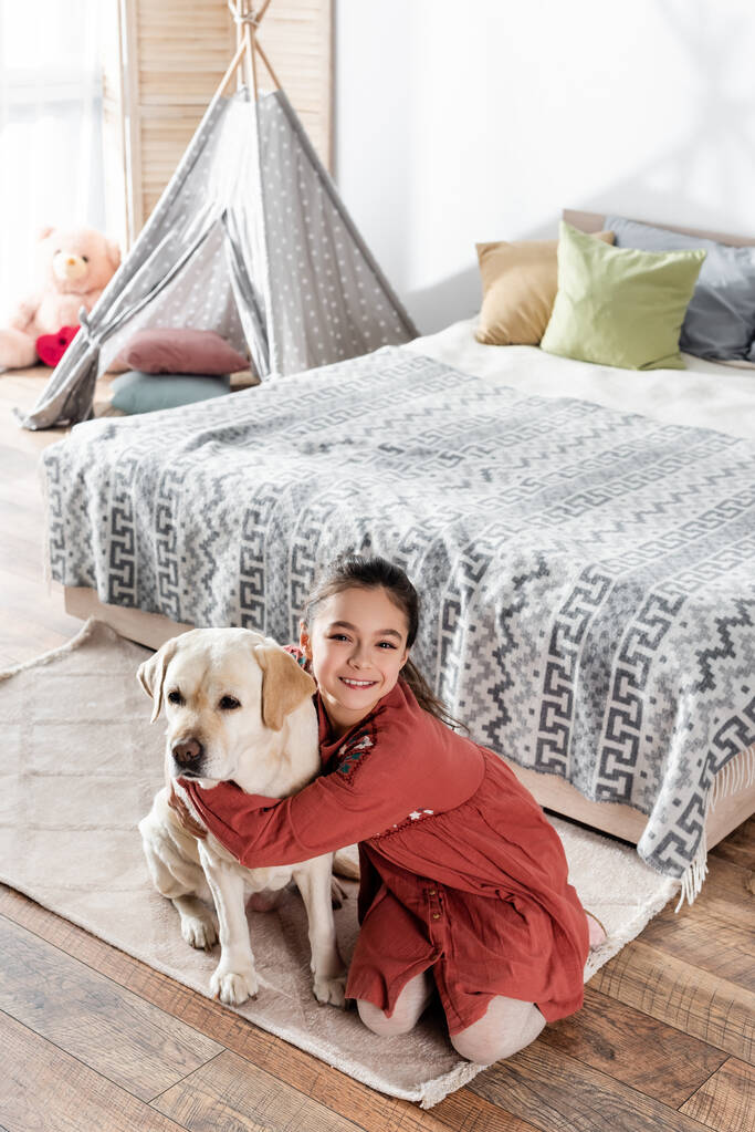 korkea kulma näkymä onnellinen tyttö halaus labrador lähellä sänkyä ja wigwam - Valokuva, kuva
