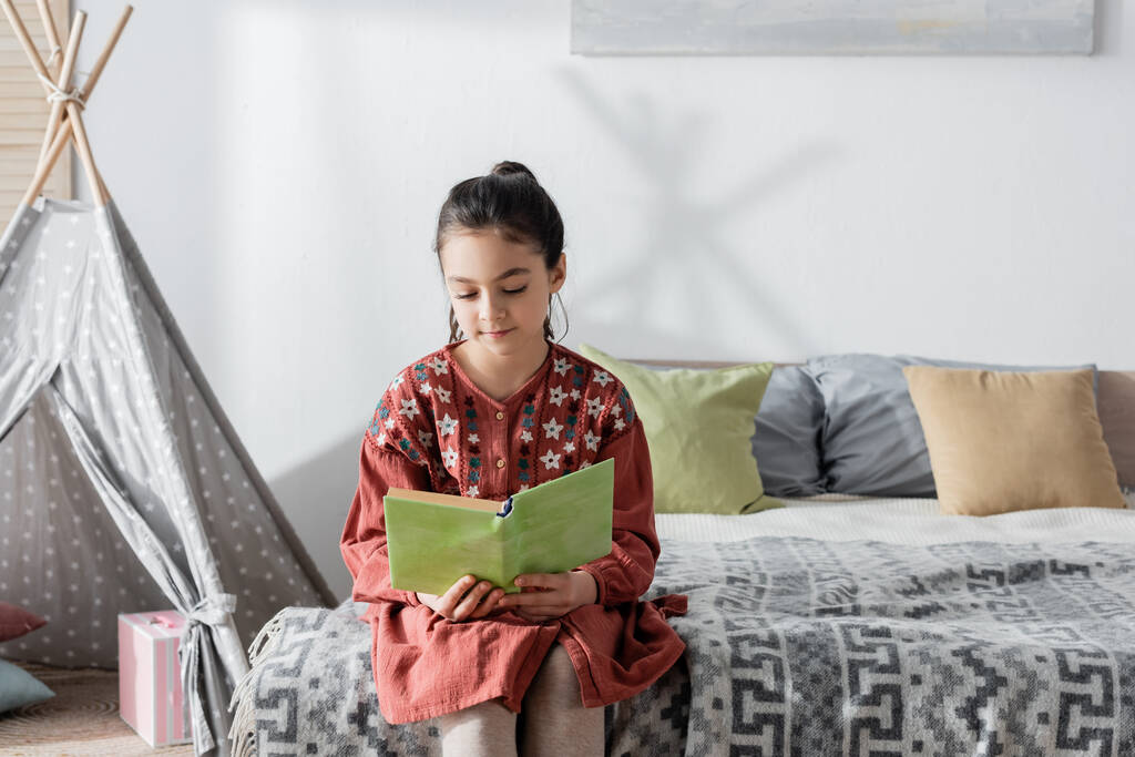 preadolescente chica leyendo libro mientras está sentado en la cama cerca de almohadas y wigwam en el fondo - Foto, Imagen