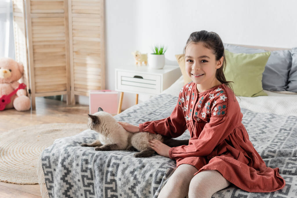 glückliches Mädchen schaut in die Kamera, während es Katze im Schlafzimmer streichelt - Foto, Bild