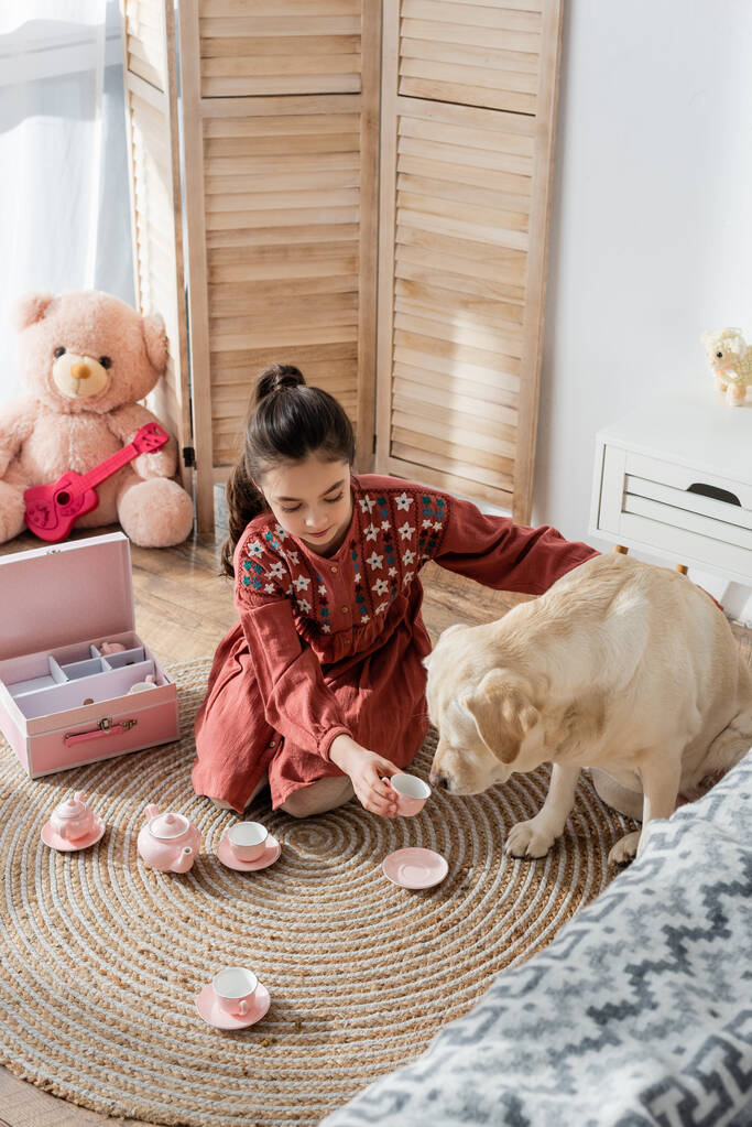 hoge hoek uitzicht op meisje spelen met speelgoed thee set en labrador hond op de vloer thuis - Foto, afbeelding
