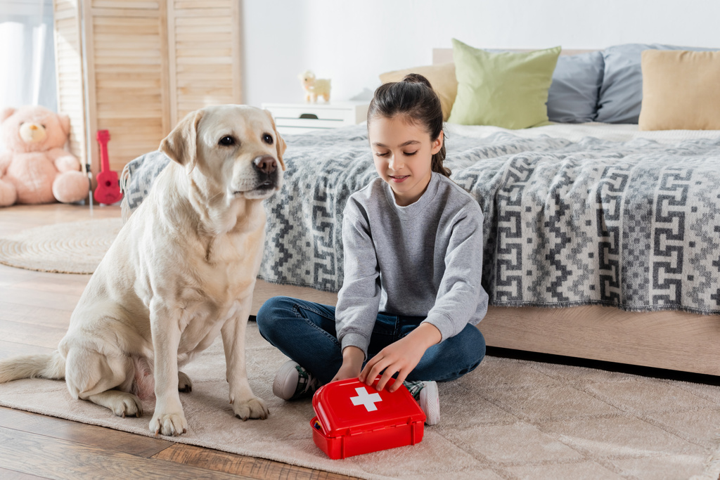 Lächelndes Mädchen spielt Arzt mit Spielzeug-Verbandskasten und Labrador-Hund auf dem Boden im Schlafzimmer - Foto, Bild