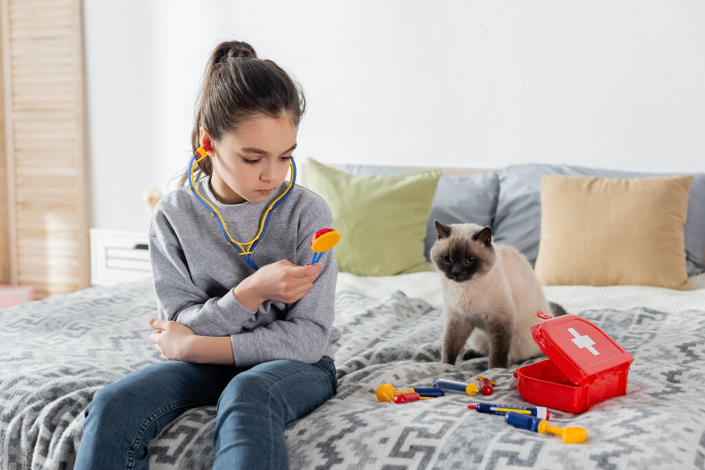 Mädchen hält Spielzeug-Stethoskop beim Spielen auf Bett neben Katze - Foto, Bild