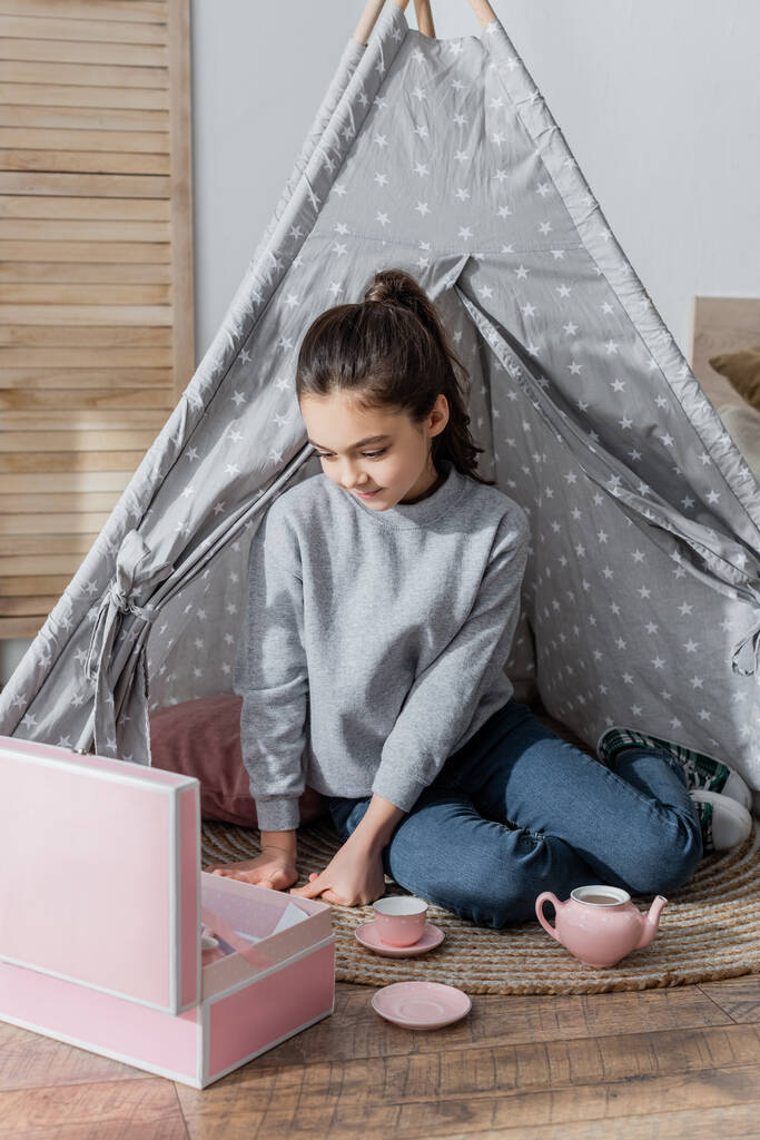 brunette preteen girl sitting on floor in wigwam and playing with toy tea set - Photo, Image