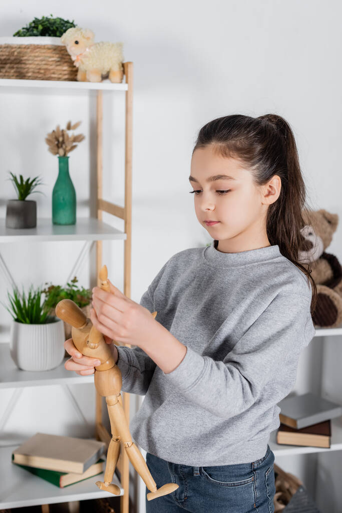 niña preadolescente sosteniendo juguete figura humana cerca de estante borroso con macetas - Foto, Imagen