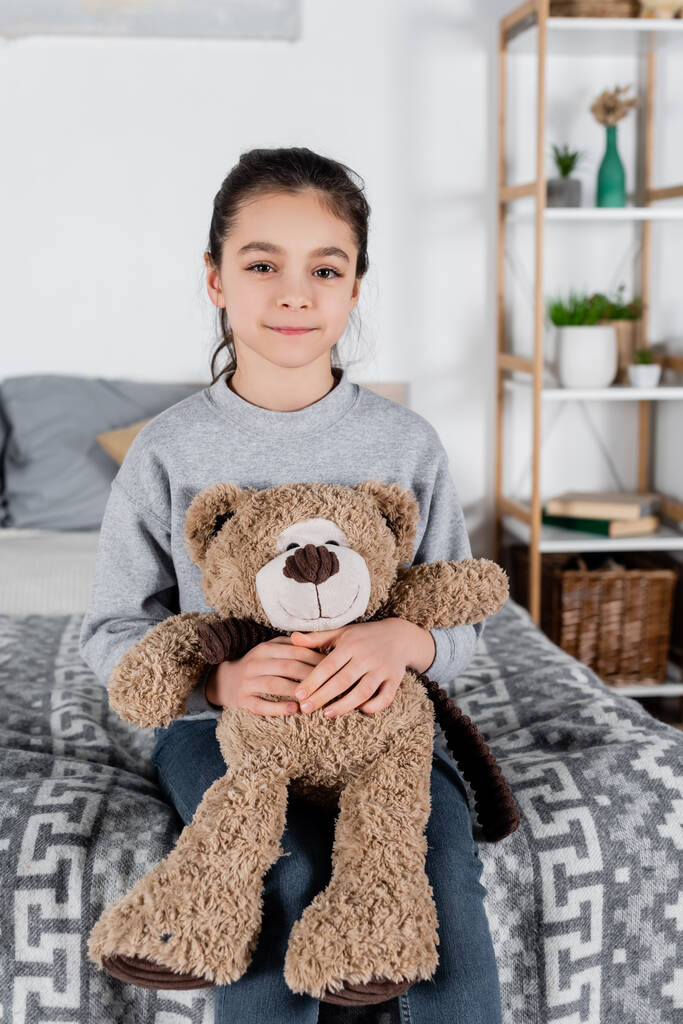 gelukkig meisje zitten op bed met teddy beer en kijken naar camera - Foto, afbeelding