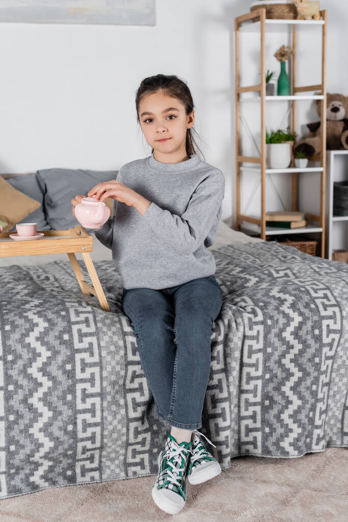 full length view of girl sitting on bed with toy teapot and looking at camera - Photo, Image