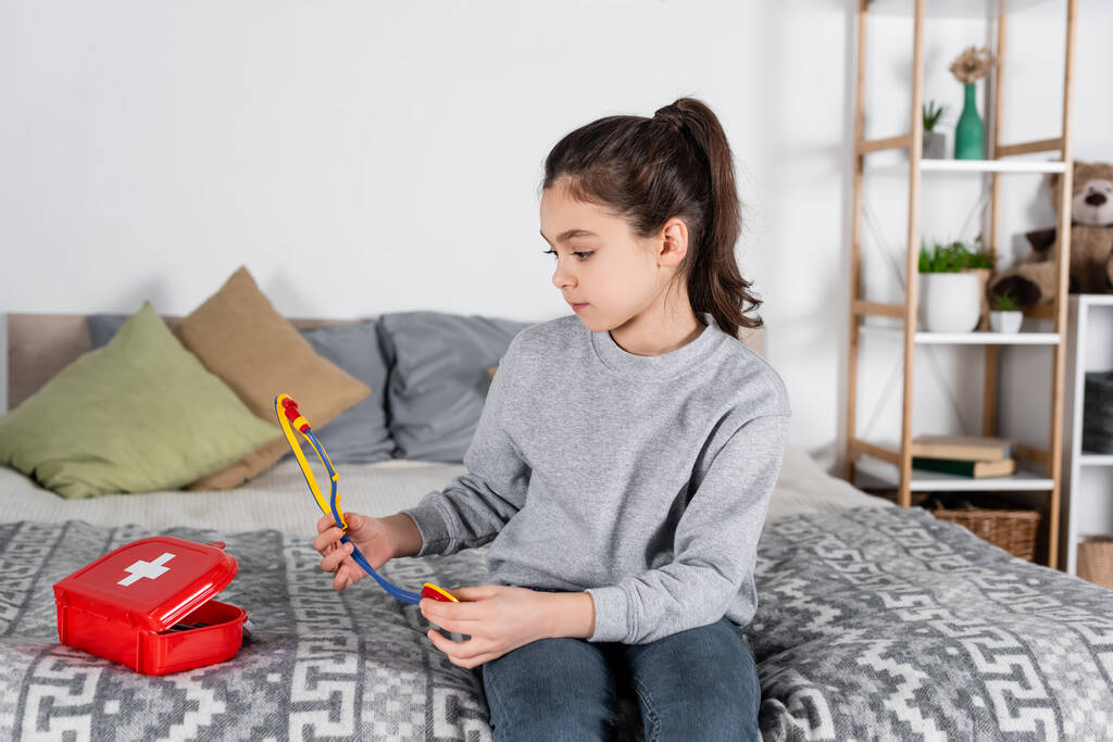 Mädchen sitzt auf Bett neben Verbandskasten und hält Spielzeug-Stethoskop - Foto, Bild