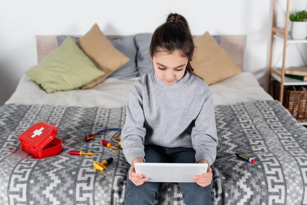 dziewczyna za pomocą tabletu cyfrowego podczas siedzenia na łóżku w pobliżu zestawu medycznego zabawki - Zdjęcie, obraz