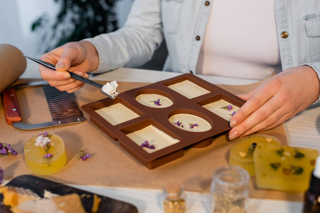 Vista cortada de artesã segurando flocos de sabão perto de molde de silicone e flores na mesa  - Foto, Imagem