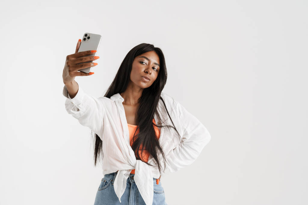 Νεαρή Αφρικανή γυναίκα με μακριά σκούρα μαλλιά που στέκεται πάνω από το λευκό φόντο του τοίχου βγάζοντας μια selfie - Φωτογραφία, εικόνα