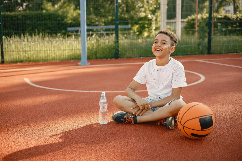 Monirotuinen poika istuu koripallokentällä vesipullon kanssa - Valokuva, kuva