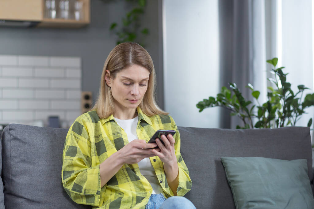 Ciddi ve odaklanmış bir kadın evinde, koltukta oturmuş, düşünceli bir şekilde telefondan haberleri okuyor. - Fotoğraf, Görsel