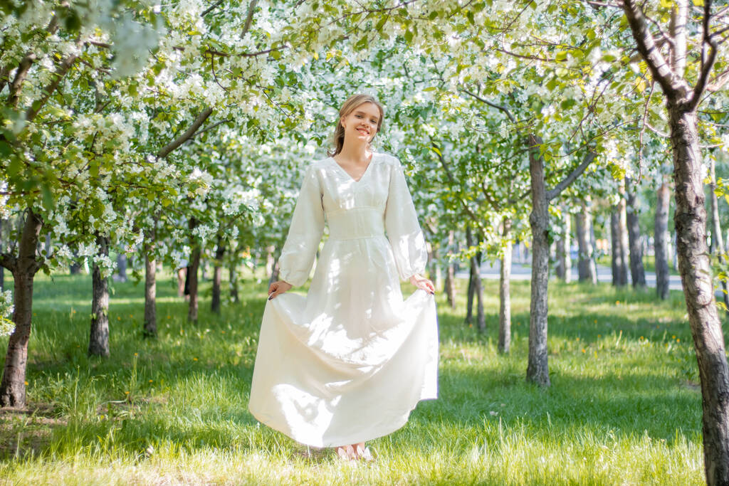 リンゴの木が咲く背景に白いドレスを着た若い女性。春の公園の花を背景にポーズをとる女の子. - 写真・画像