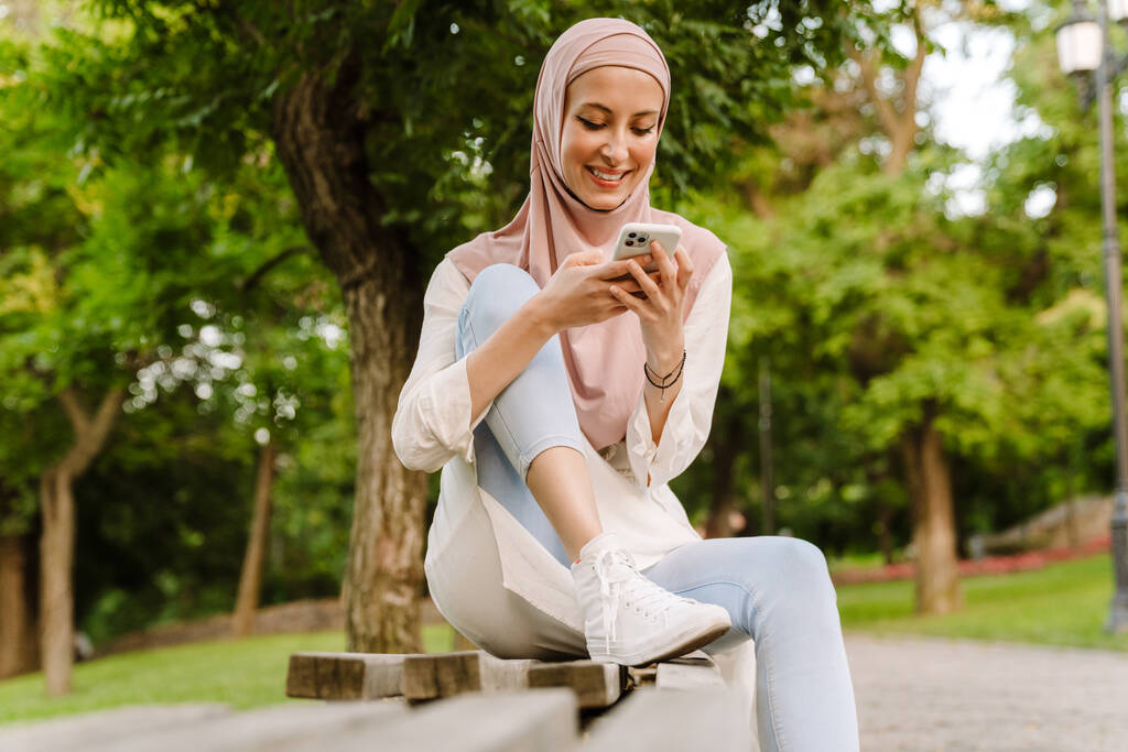 Smiling middle aged muslim woman sitting on a bench outdoors using mobile phone - Photo, Image