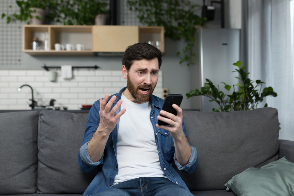Muž doma, šokovaný a zklamaný, se podívá na telefon, sedící na gauči - Fotografie, Obrázek