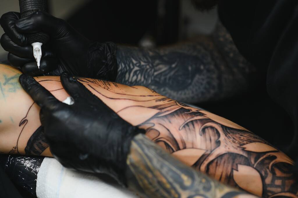 Gros plan d'un tatoueur barbu travaillant dans son atelier tatouant sa manche sur le bras de son client masculin. Homme se faire tatouer par tatoueur professionnel
 - Photo, image